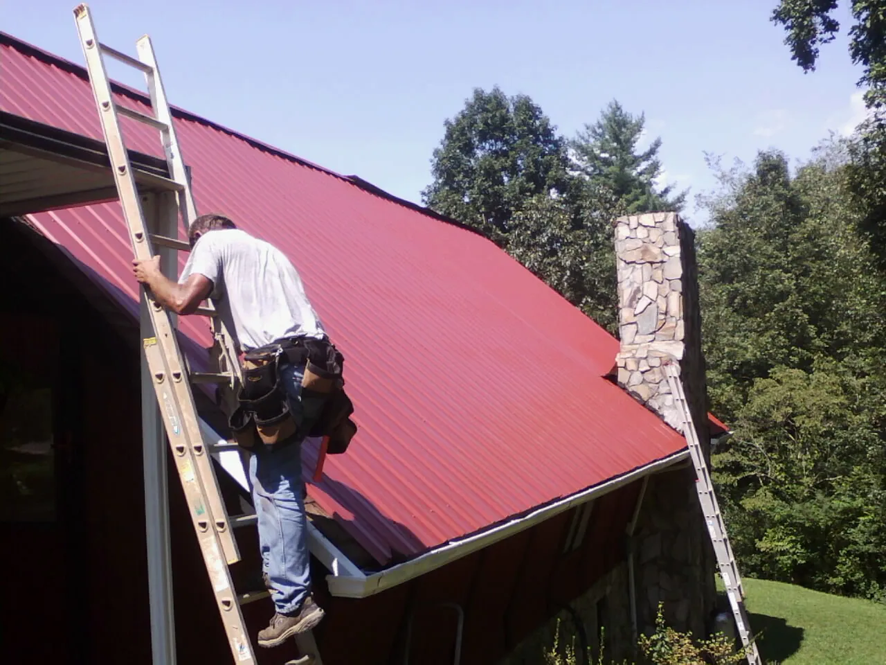 Roofer on a ladder beside a red metal roof and stone chimney — Soffit & Fascia Repair by Roofing Brothers in Shellsburg
